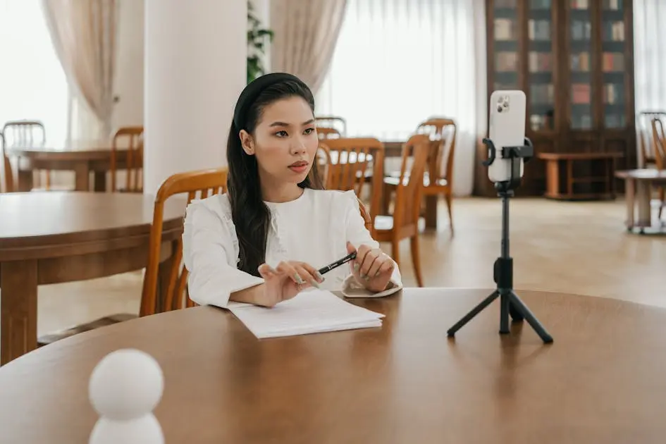 An independent asian woman sitting at a table with documents, looking seriously at her smartphone on a tripod, representing a single woman in malaysia proactively managing her estate planning.