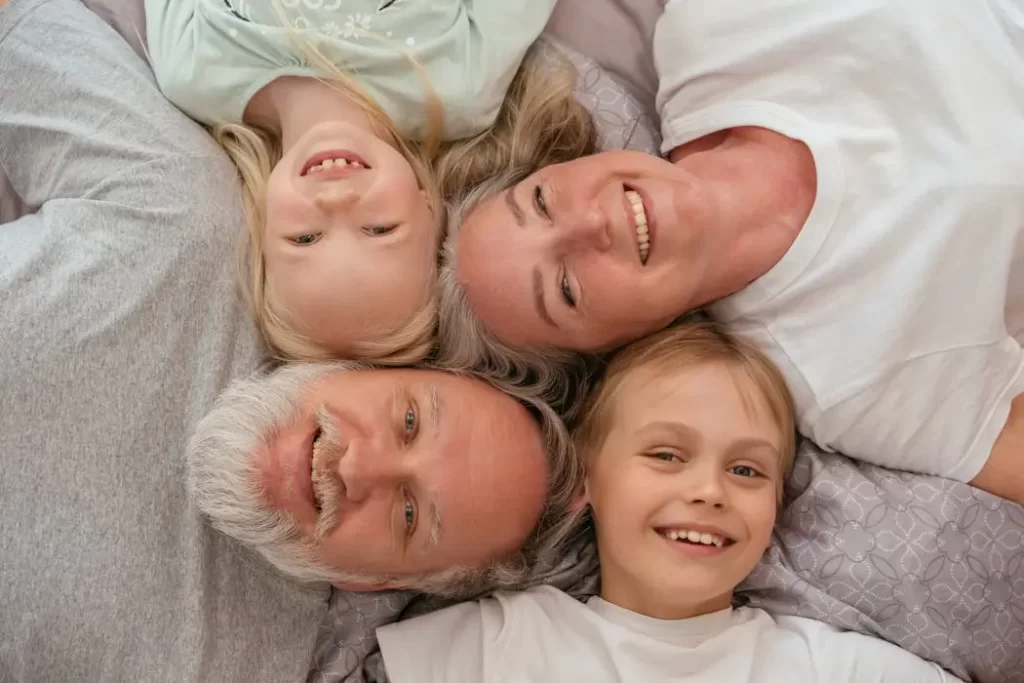 An older couple and two young children lying closely together on a bed and smiling, representing a happy blended family that requires careful estate planning in malaysia.