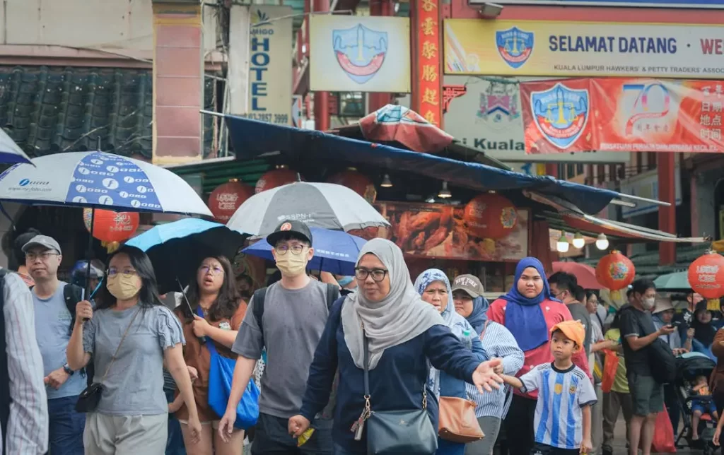 A crowded street scene in kuala lumpur with pedestrians walking in the rain, symbolizing the everyday malaysians who are unaware of the billions held by the registrar of unclaimed moneys.