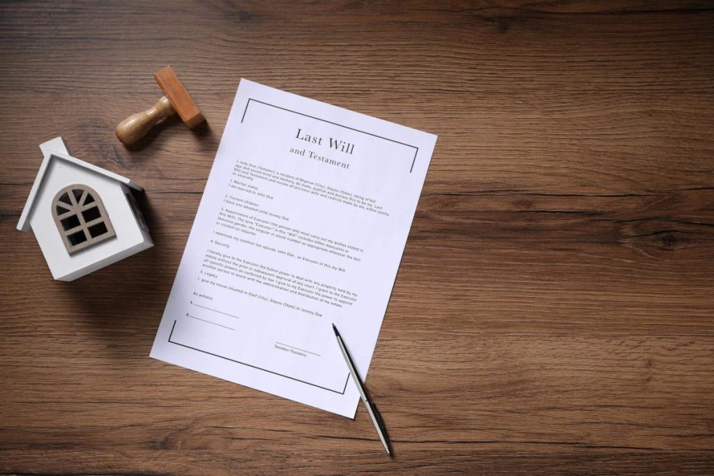Sheet titled 'last will and testament' on a wooden desk, with a tiny white house model, a stamp, and a pen nearby.